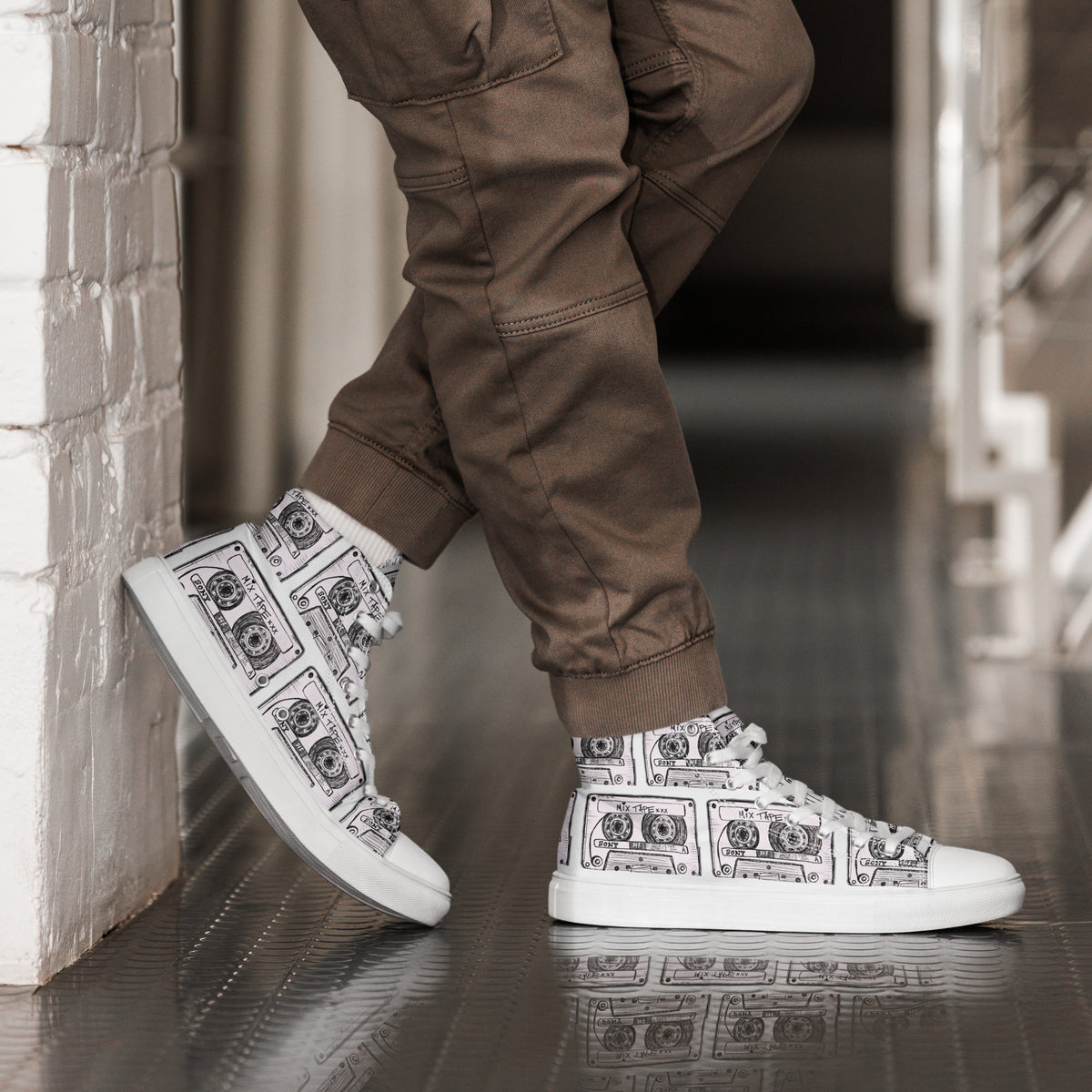 A person wearing Vicky Steane's Mix Tape white high-top sneakers  standing on a reflective dark floor next to a white brick wall.